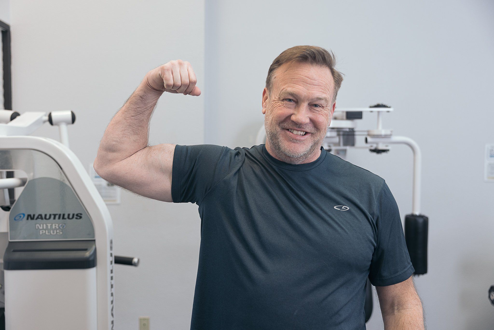 Member proudly flexing his arm muscles after a strength training session at The Perfect Workout gym with Nautilus equipment in the background.