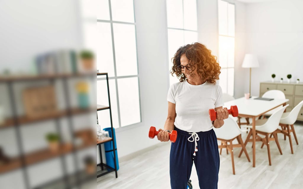 Middle-aged woman with curly hair lifting weights at home, focused on maintaining her fitness routine