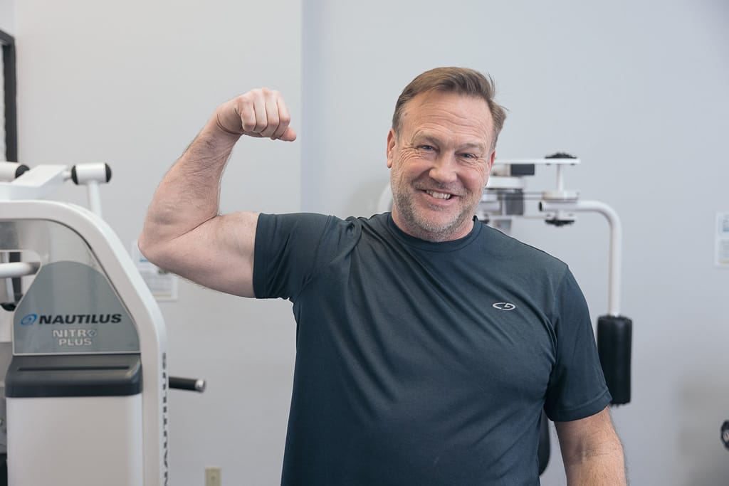Member proudly flexing his arm muscles after a strength training session at The Perfect Workout gym with Nautilus equipment in the background.