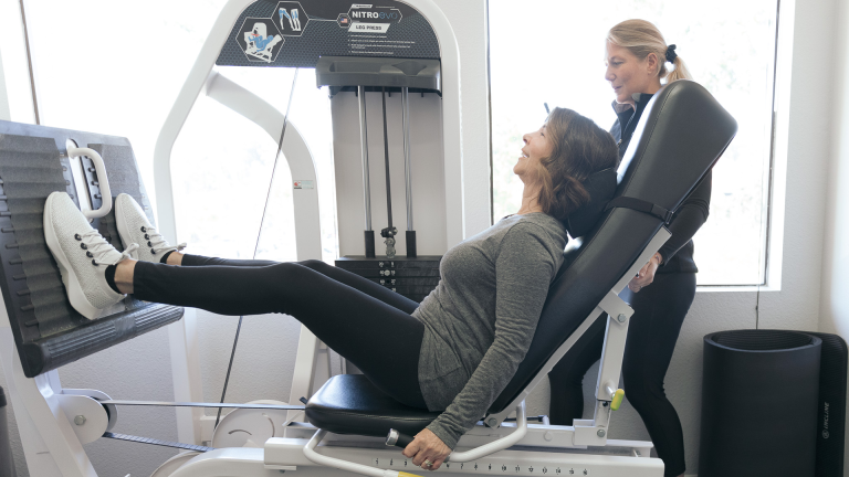 A trainer guides a woman on the leg press machine at The Perfect Workout